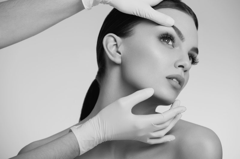 Black and white studio photograph of a woman's face held between two gloved hands, one resting at the brow and one under the jaw.