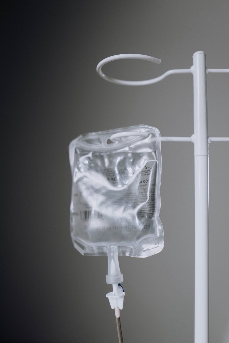 A clear fluid bag hanging from a white drip stand against a plain grey wall, the line running down out of frame.