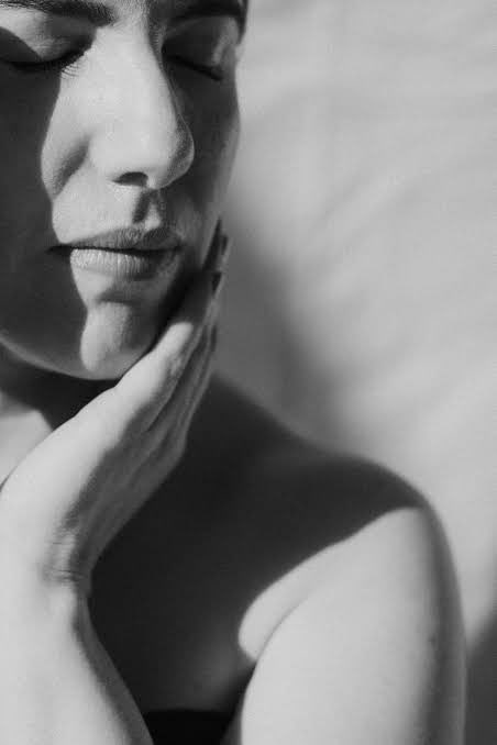 Black and white photograph in soft daylight of a woman with her eyes closed and the back of her hand resting against her cheek.