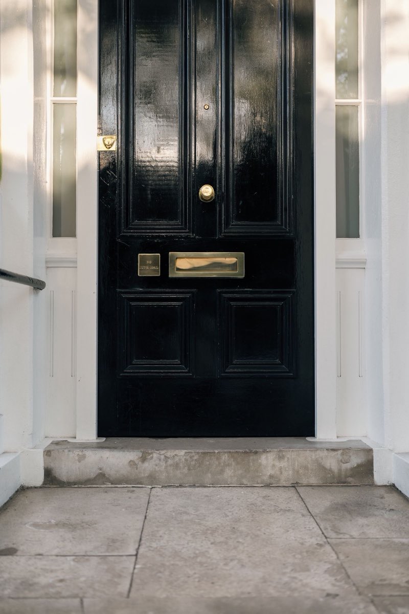 A gloss black panelled front door with a brass knob and letterplate, set in a white painted surround above a worn stone step.